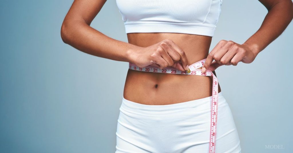 A woman wearing a white bra and underwear is holding a measure tape around her waist. (model)